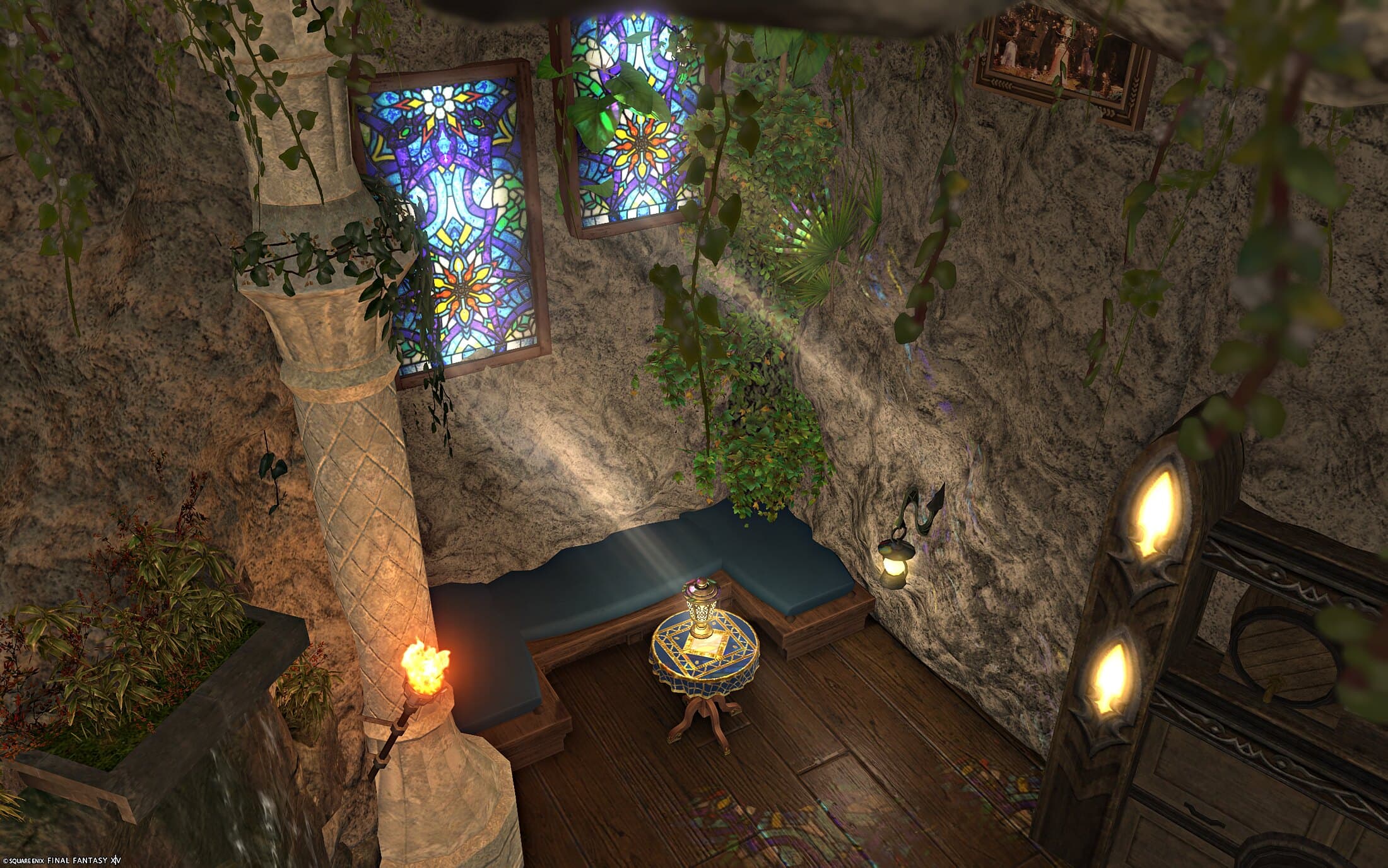 The seating nook in the tavern, from above — a blue cushioned bench around a corner and a small round blue-topped table with a lantern, under two stained-glass windows set in the rough stone wall, light falling from them, an ivory pillar wound with ivy and a burning torch at the left