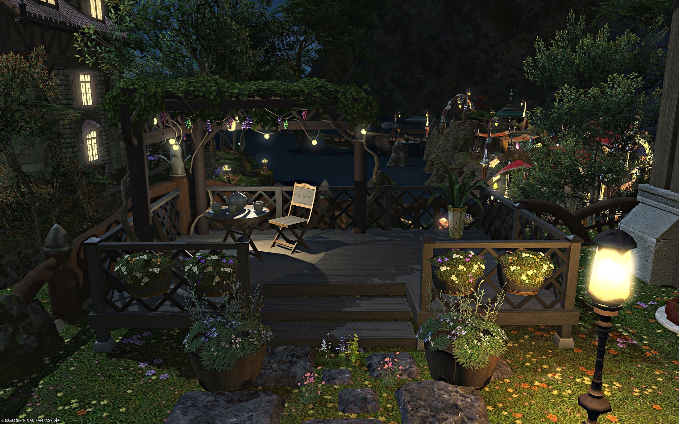 A vine-covered deck with a café table and chairs, string lights overhead, and a lake and lantern-lit rockwork beyond the railing