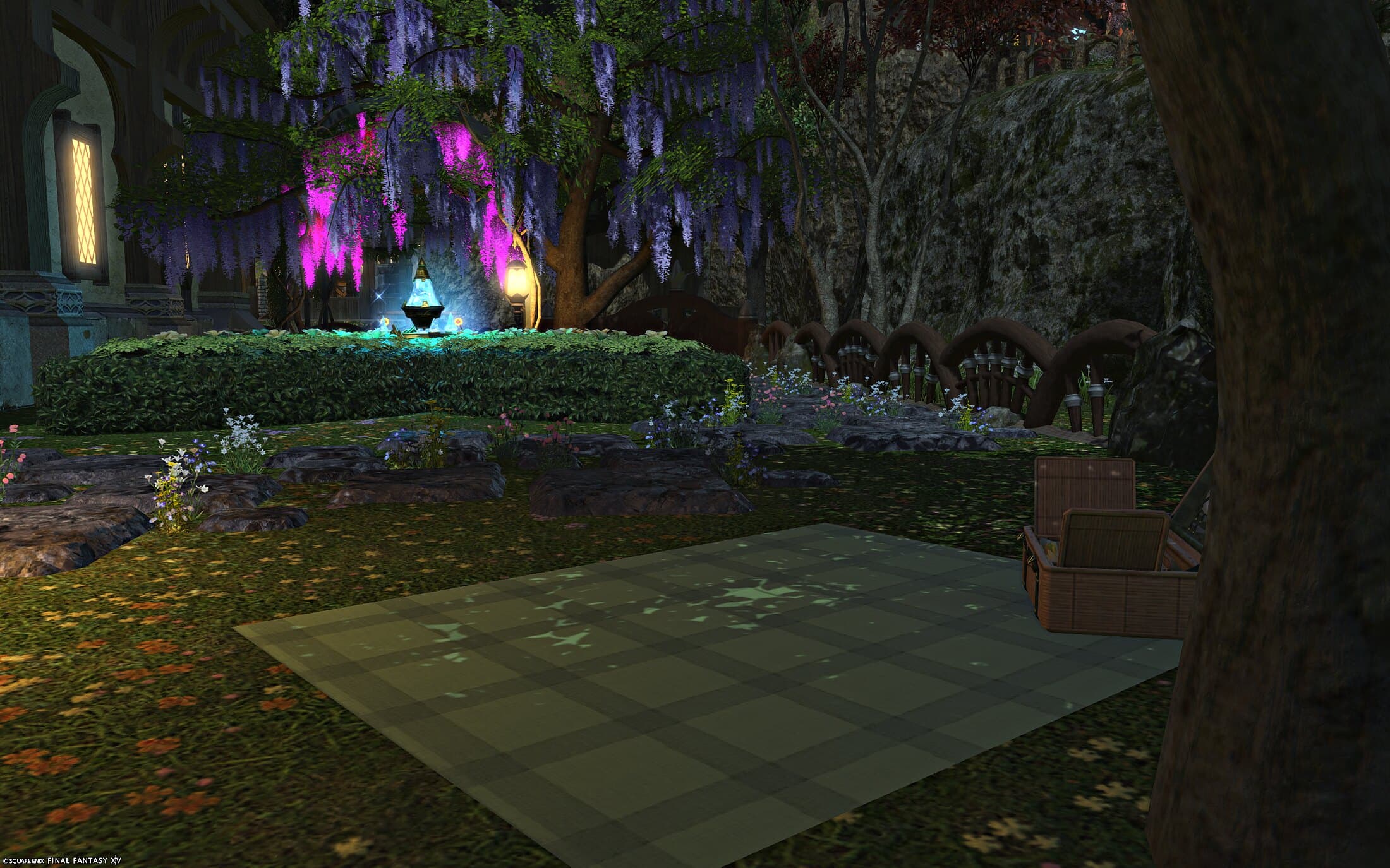 The garden hedge and its glowing fountain seen past a picnic basket and a rug laid out on the lawn